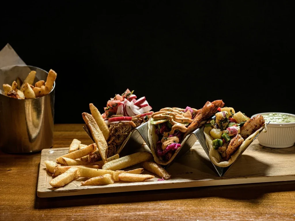 Three gourmet tacos, french fries, and green dip on a wooden board.