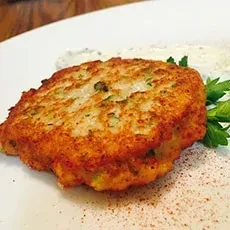 Crispy, golden-brown fried patty on white plate with herbs and sauce.