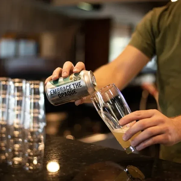 Close-up of hands pouring amber drink from can labeled "EMBRACE THE OPAQUE."