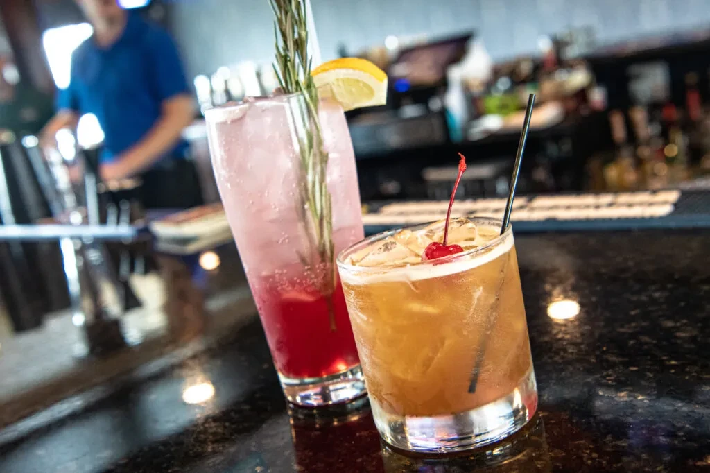 Two cocktails, amber and pink, on a bar with a blurred bartender.