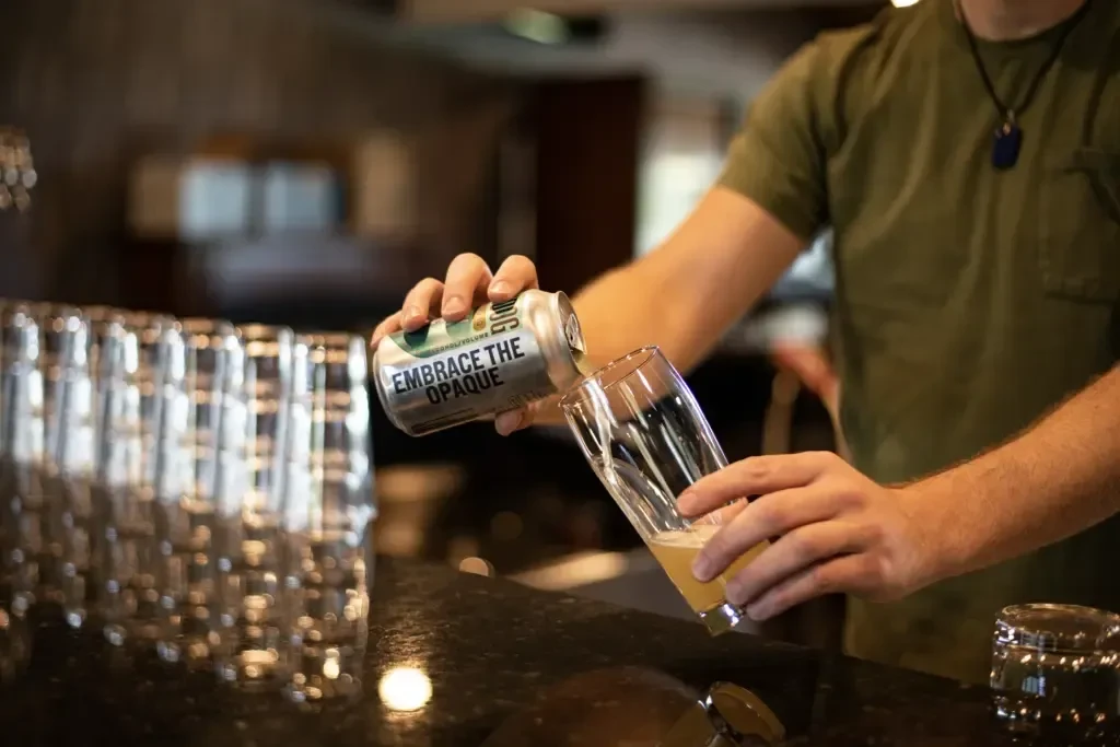Hands pour opaque beer from silver can into clear glass.