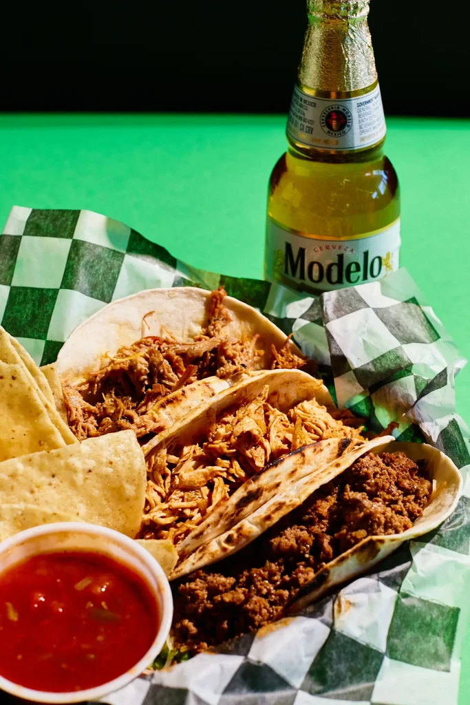 Three tacos, chips, salsa, and Modelo beer on a green surface.