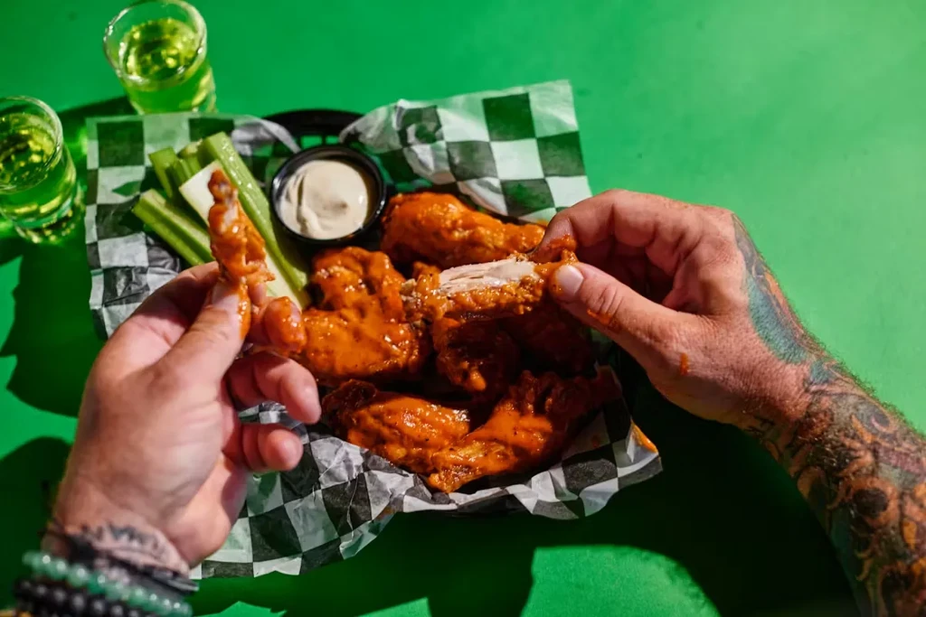 Hands eating saucy buffalo chicken wings, celery, and dipping sauce.