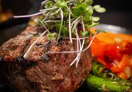 Close-up of a grilled steak plated with microgreens, asparagus, and bell peppers.