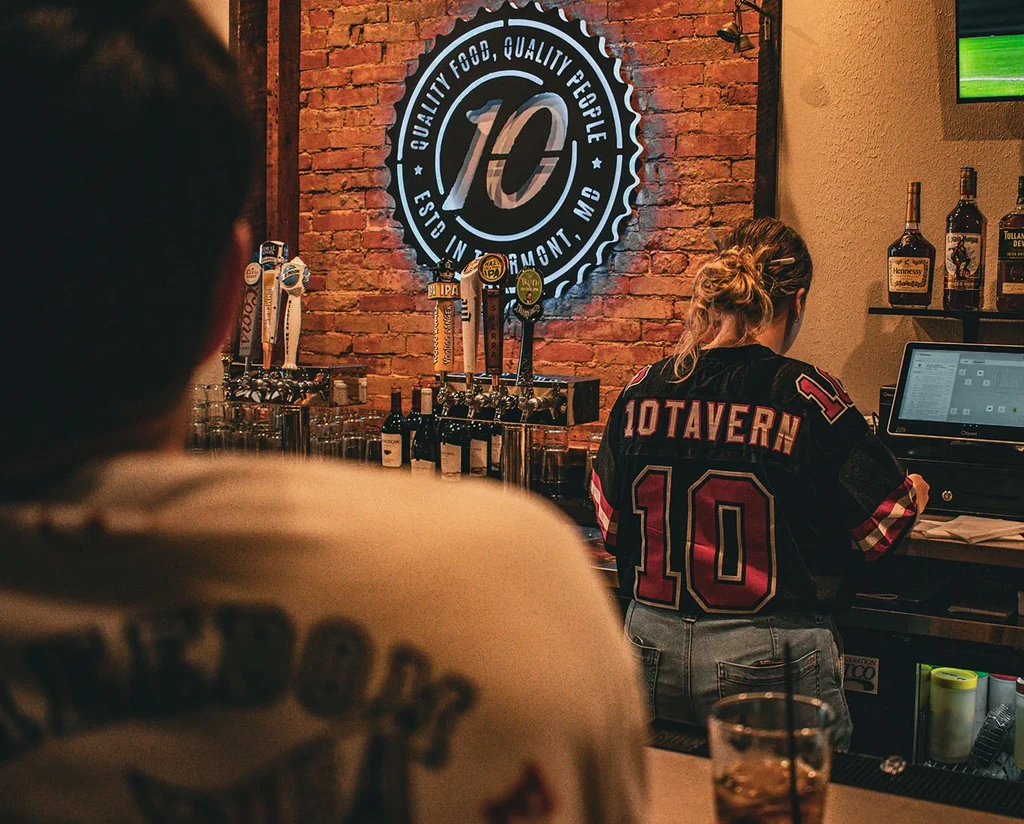 Dim bar with '10' neon sign, taps, and bartender.