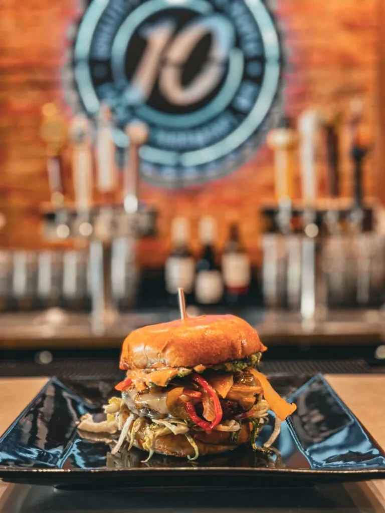 Loaded gourmet burger on dark plate, blurred bar with "10" logo.