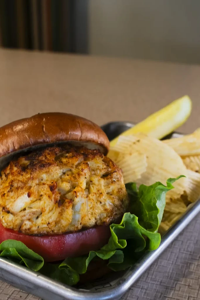 Crab cake sandwich, chips, and pickle spear on metal tray.