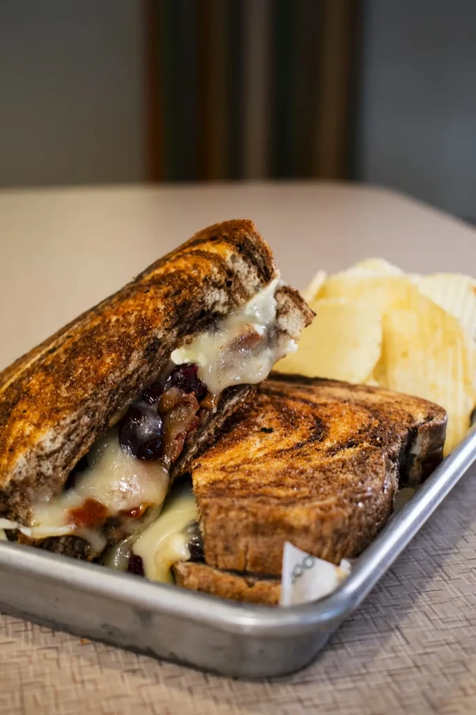 Grilled sandwich with oozing cheese, dark filling, and crinkle-cut chips.