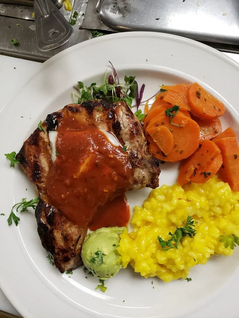 Plated grilled meat, orange carrots, yellow risotto, and herb garnish.