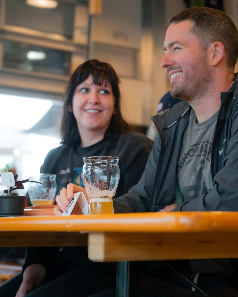Smiling man and woman holding beer at a brewery table.