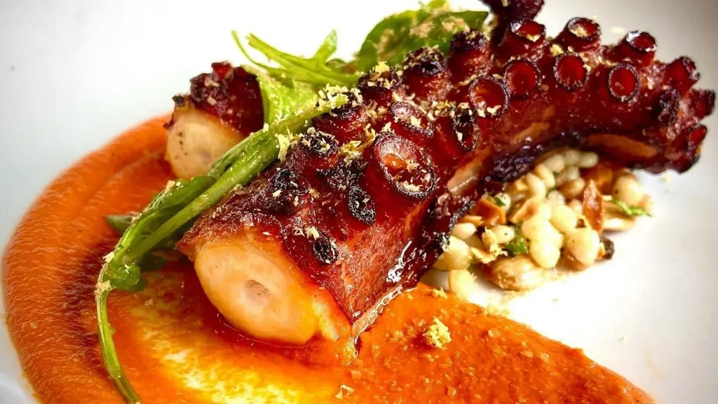 Close-up of grilled octopus tentacle, orange sauce, white beans, green herbs.