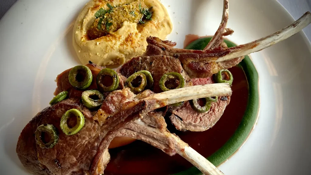 Plated lamb chops with green olives, purée, sauce, and hummus.