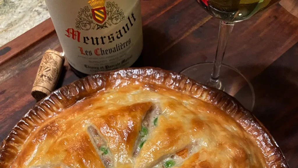 Golden-crusted pie, Meursault wine bottle, and glass on a wooden board.