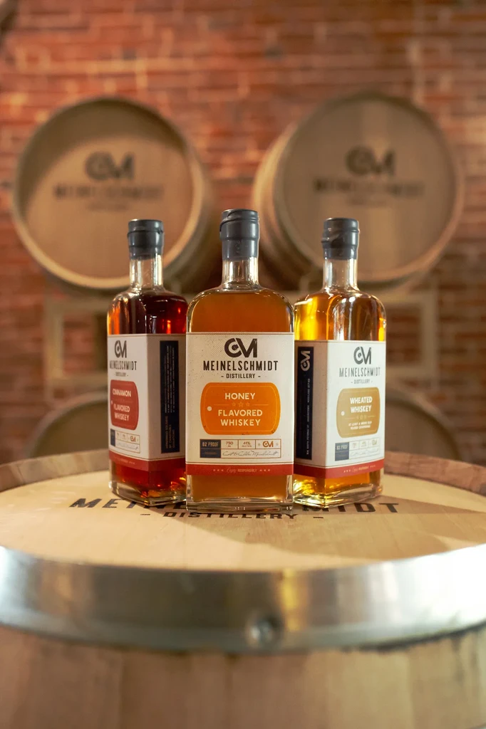 Three Meinelschmidt whiskey bottles (cinnamon, honey, wheated) on a wooden barrel.