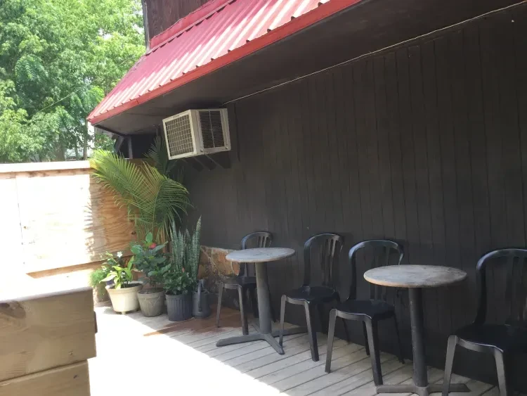 Sunny outdoor patio: dark wooden wall, red roof, potted plants, tables.