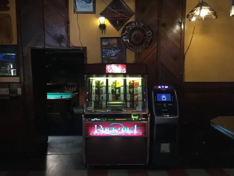 Lit-up Rock-Ola jukebox and ATM in a dimly lit, rustic bar.