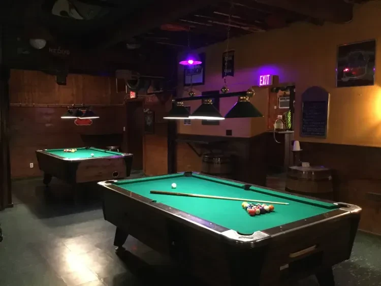 Dimly lit bar interior with two pool tables, pendant and neon lights.