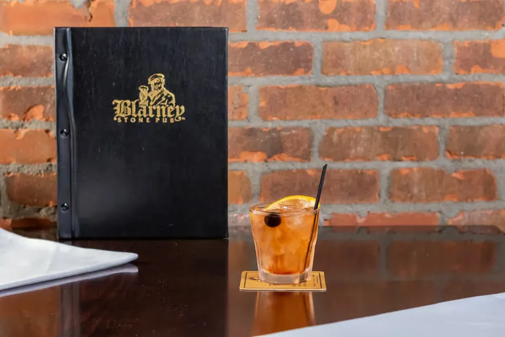 Amber cocktail and 'Blarney Stone Pub' menu against a brick wall.