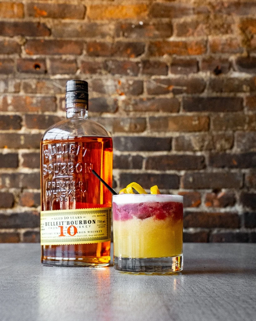 Bulleit Bourbon bottle and colorful layered cocktail with lemon peel.
