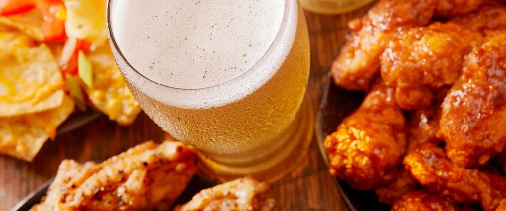 A frosty beer, glazed wings, and nachos on a wooden table.