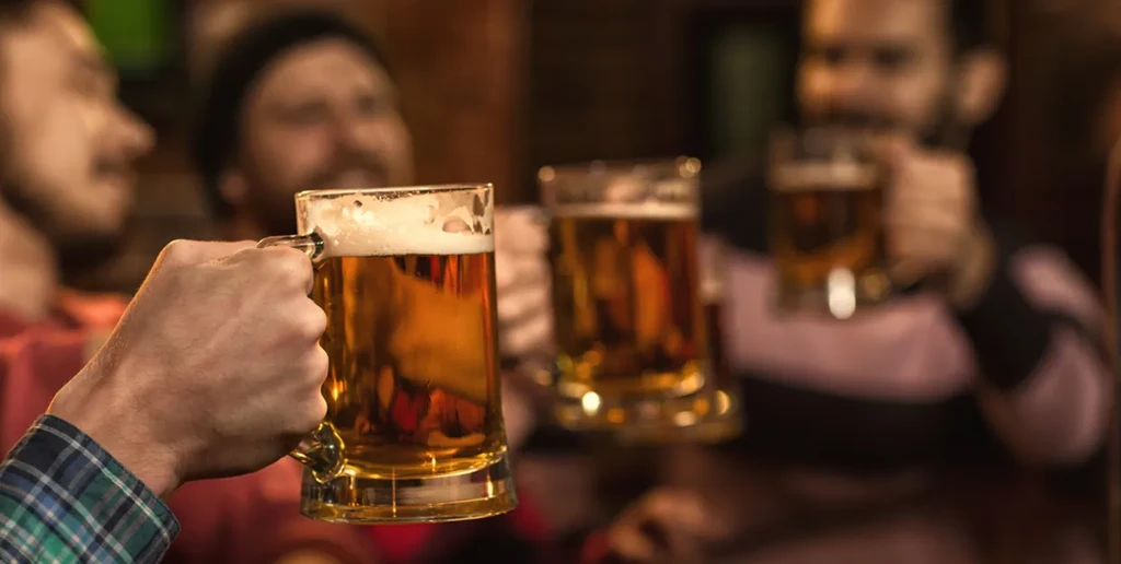 Male hand holding a frothy beer mug, toasting with blurred friends.