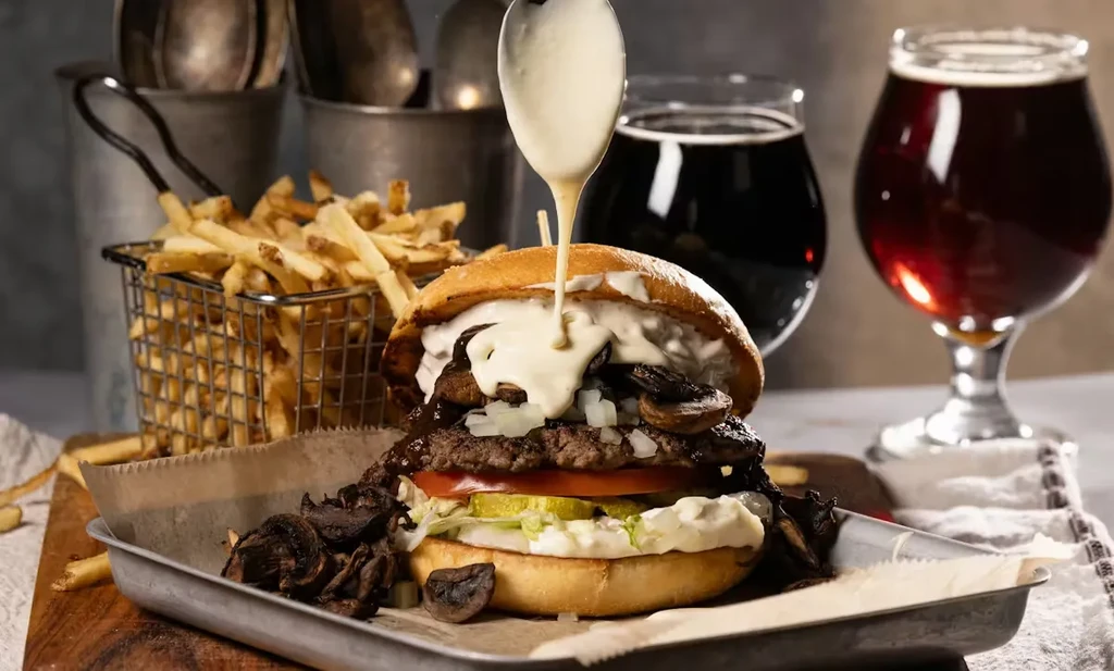 Gourmet burger drizzled with white sauce, fries, and beer on tray.