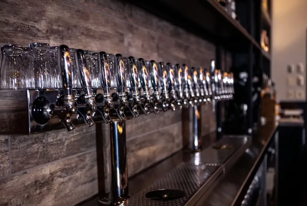 Shiny chrome beer taps and inverted glasses on a rustic bar wall.