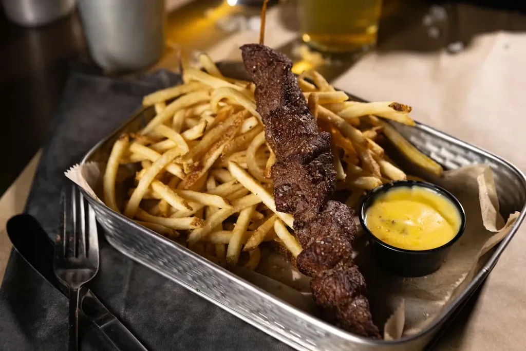 Golden french fries, grilled meat skewer, and yellow dipping sauce in a tray.