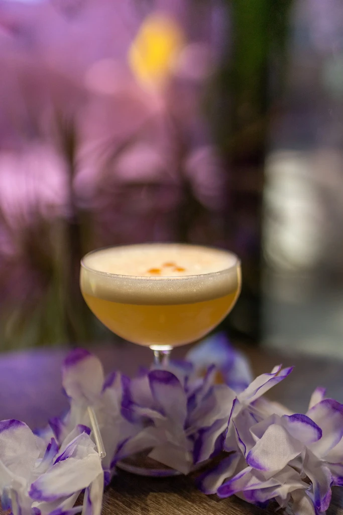 Yellow-orange cocktail, white foam, floral lei, blurred colorful lights.