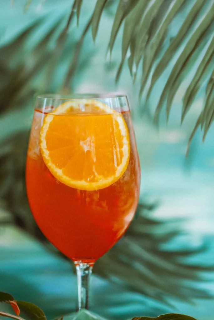 Orange cocktail in stemmed glass with orange garnish and blurred tropical background.