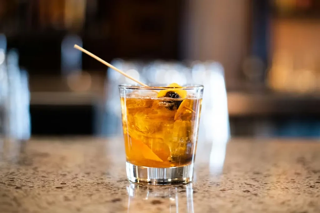 Amber cocktail with ice, orange twist, and cherry in a short glass.