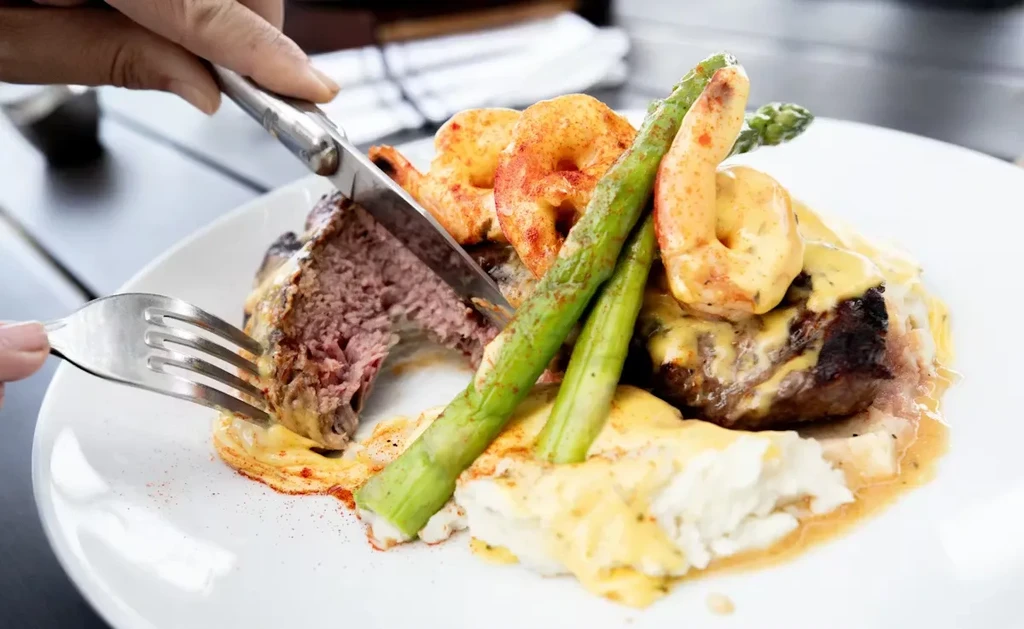 Hands cut into medium-rare steak, shrimp, asparagus, mashed potatoes, with sauce.