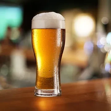 A cold pilsner glass filled with golden beer and frothy foam on wood.