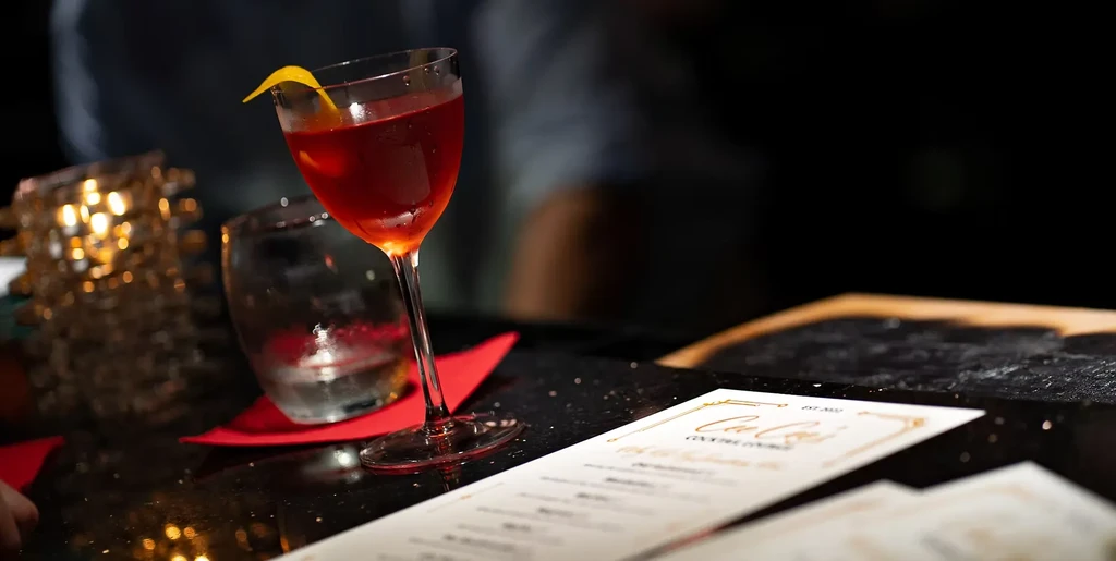 Deep red cocktail with lemon peel, menu, and warm bokeh lights.