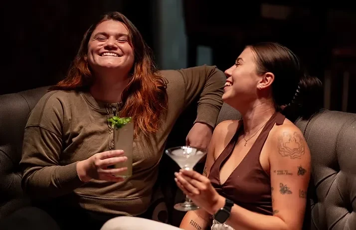 Two women laughing, holding drinks in a dimly lit bar booth.