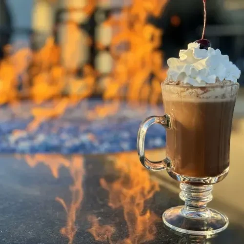 Glass mug with dark drink, whipped cream, cherry, by fire pit.
