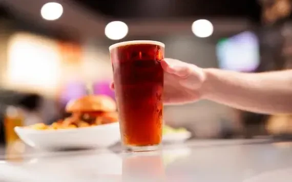 Hand holding a glass of beer; burger and side dish behind.
