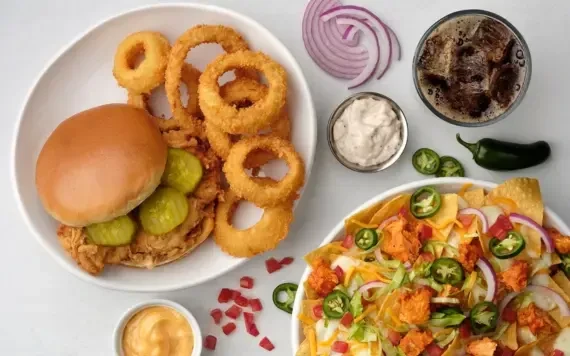 Flat lay of fried chicken sandwich, loaded nachos, sauces, and drink.