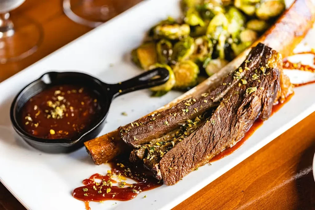 Slow-cooked meat on bone with dark sauce, herbs, and green vegetables.