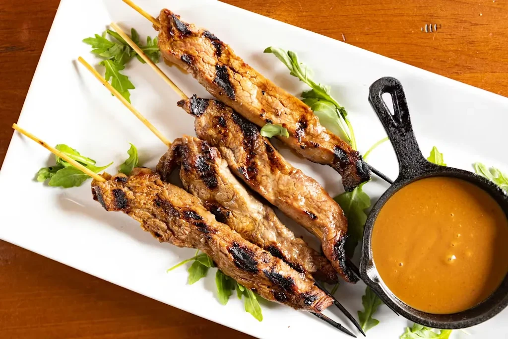 Five char-grilled meat skewers, green garnish, and creamy dipping sauce.