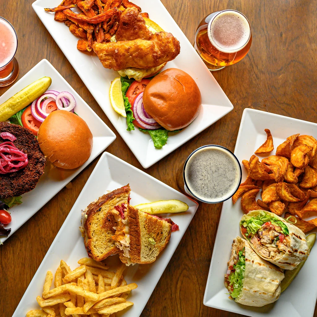 Flat lay of diverse restaurant food and drinks on a warm wooden table.