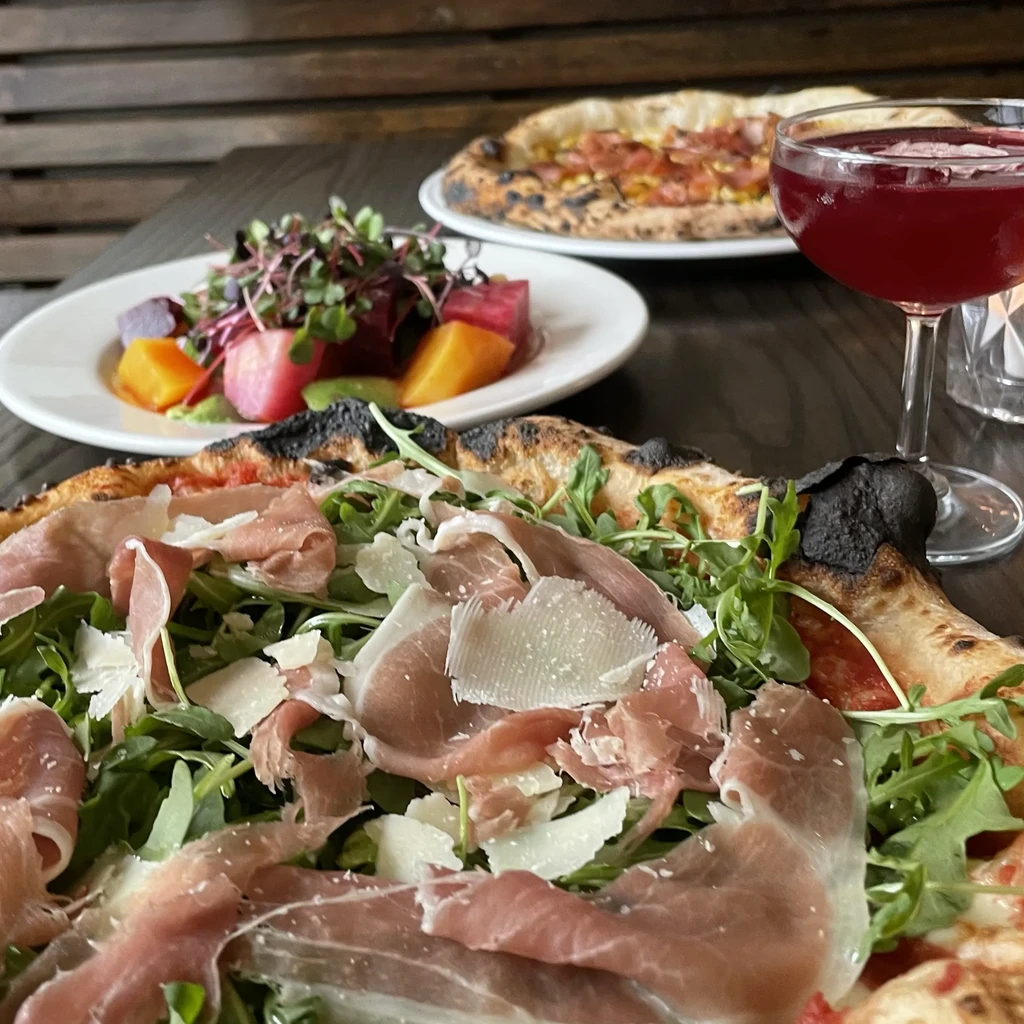 Prosciutto pizza, beet salad, and dark red drink on a wooden table.