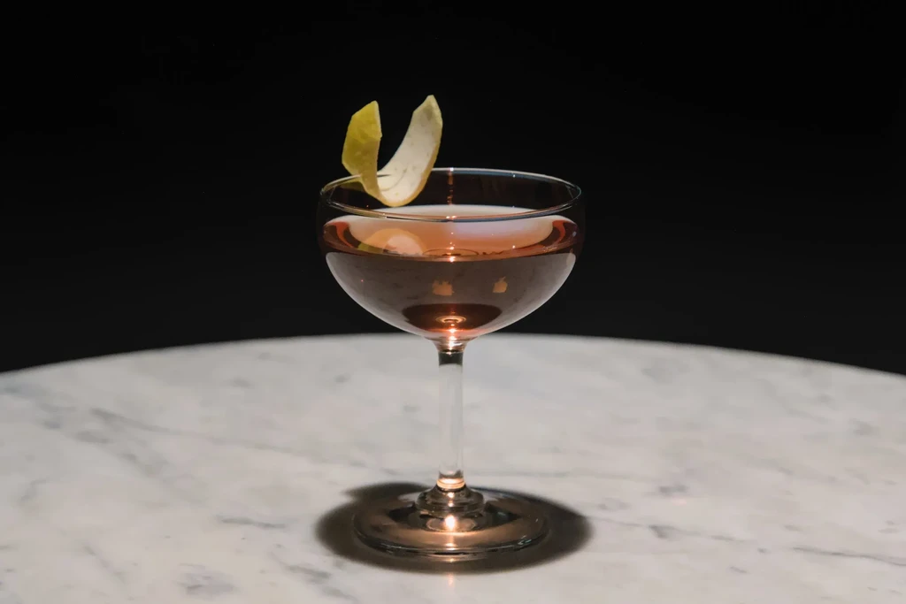 Reddish-brown cocktail in coupe glass with twisted citrus peel on marble.