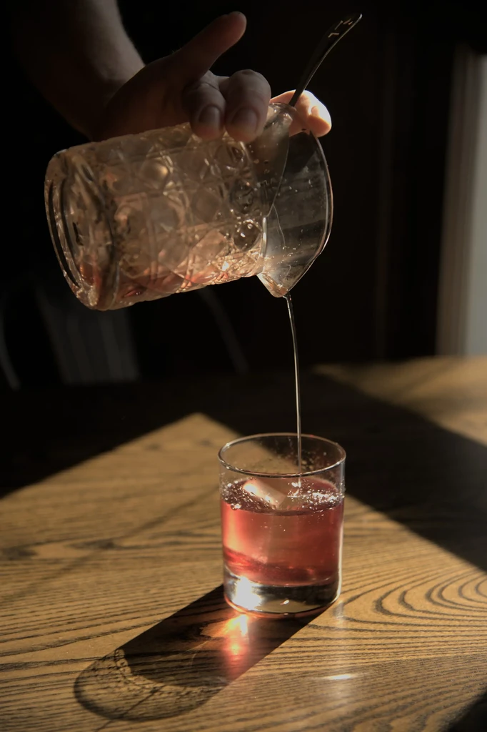 Hand pouring a red drink from mixing glass into rocks glass.