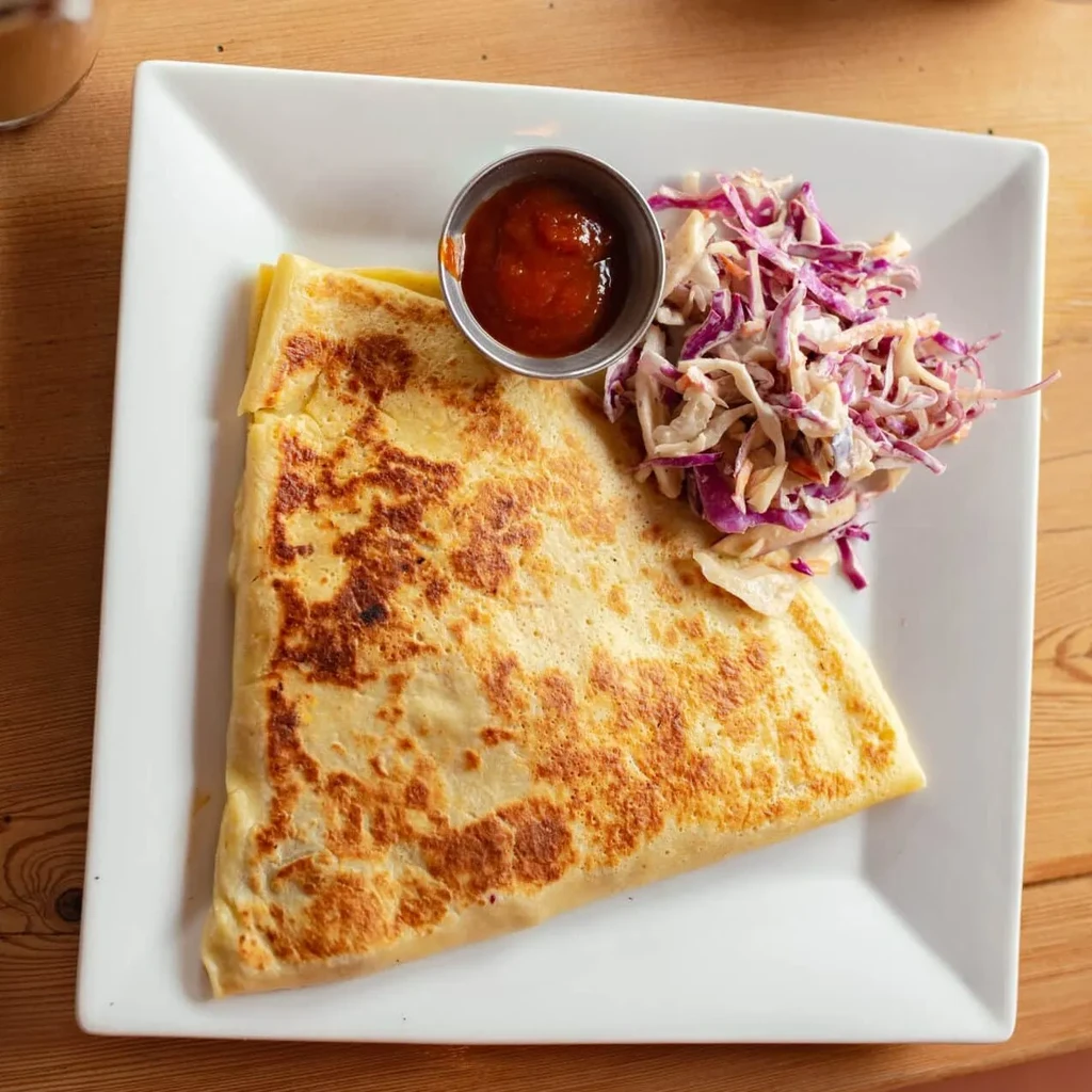 Plate with golden-brown folded crepe, coleslaw, and red sauce.