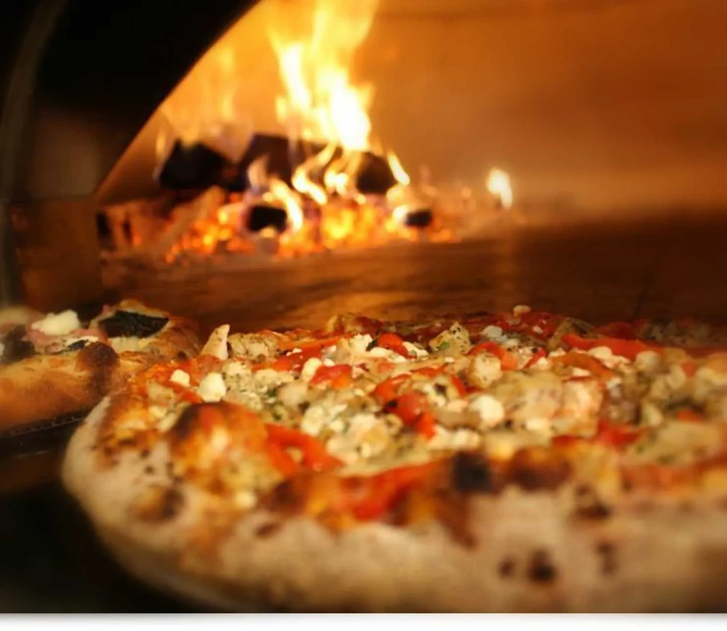 Two pizzas cooking in a wood-fired oven with visible flames.