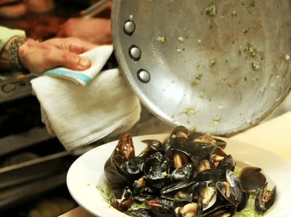 Mussels in green sauce steaming on a plate, hand holds cooking pan.