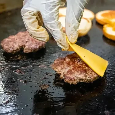 Gloved hand places yellow cheese on sizzling burger patty on a griddle.