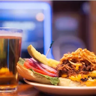 A pulled meat burger, pickle spear, and beer on a wooden surface.