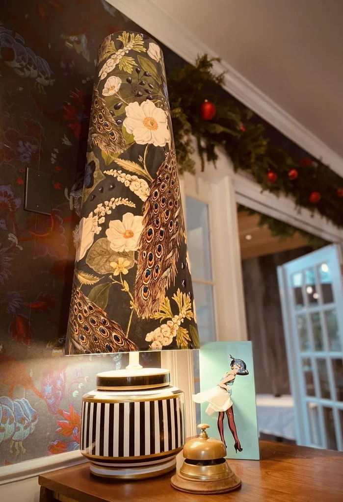 Decorative lamp with peacock feather shade, striped base, brass bell, and festive garland.